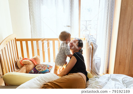 Smiling mom lies on the bed near the standing baby on the background of a crib and toys Smiling mom lies on the bed near the standing baby on the background of a crib and toys 79192189