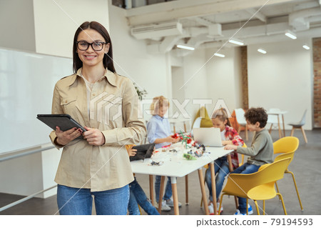 Beautiful young female teacher in glasses smiling and holding tablet pc while posing for camera. Kids working on diy robot in the background Beautiful young female teacher in glasses smiling and holding tablet pc while posing for camera. Kids working on diy robot in the background 79194593