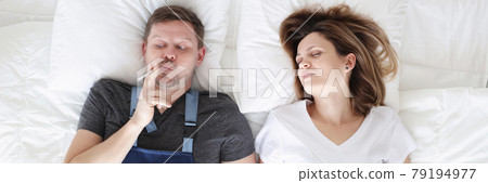 Smoking man with cigarette lies in bed with his sleeping wife Smoking man with cigarette lies in bed with his sleeping wife 79194977
