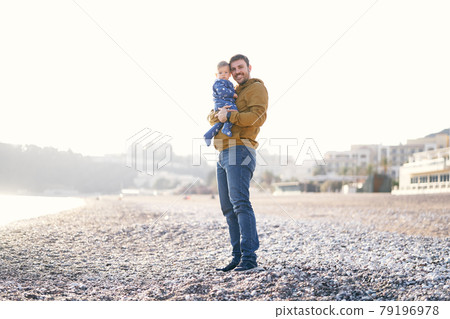 Smiling dad in a brown jacket and jeans holds a small child in a blue overalls in his arms while standing on a pebble beach Smiling dad in a brown jacket and jeans holds a small child in a blue overalls in his arms while standing on a pebble beach 79196978