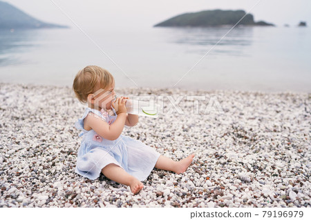 Little girl in a dress drinks water from a bottle on a pebble beach 79196979