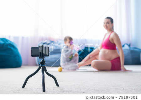 A pregnant woman does gymnastics with her son live. The expectant mother shoots a training video for her blog on the phone. Fitness with a baby. 79197501