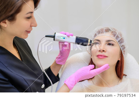 Portrait of a middle-aged woman receiving a non-surgical rejuvenation procedure at a cosmetologist. RF face lifting. Hardware facial massage. 79197534