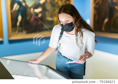 Young girl in a protective mask in the museum looks at the exposition behind a glass case Young girl in a protective mask in the museum looks at the exposition behind a glass case 79198821