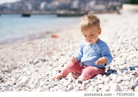 Pensive kid sits on a pebble beach, looking at his feet, near the sea 79199165