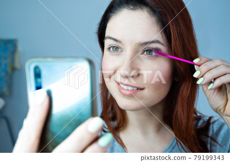 Beautiful redhead woman paints eyelashes and takes a selfie. The girl puts mascara and looks at the phone instead of a mirror. 79199343