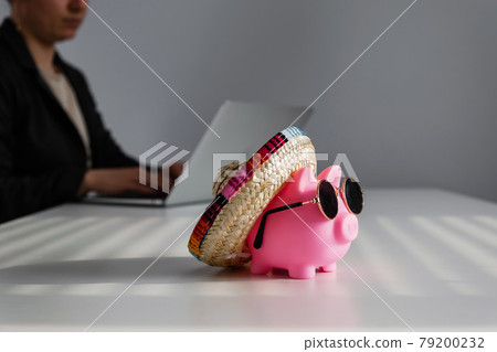 Piggy bank in sunglasses and a sombrero in the background woman works for a laptop 79200232