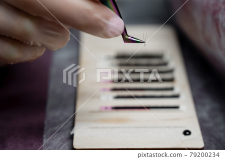 Close-up. The master takes a bunch of curved artificial eyelashes with tweezers from the tablet and dips into the glue. The process of eyelash extension in a beauty salon. Rows of material. 79200234