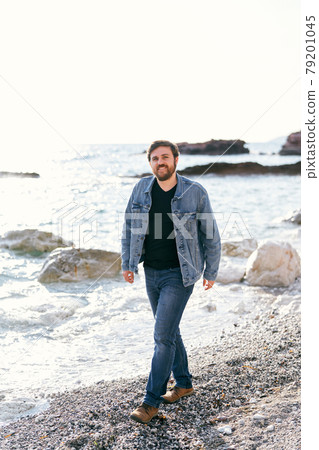 Smiling man in jeans and a denim jacket walks along the pebble beach near the water against the backdrop of stones Smiling man in jeans and a denim jacket walks along the pebble beach near the water against the backdrop of stones 79201045