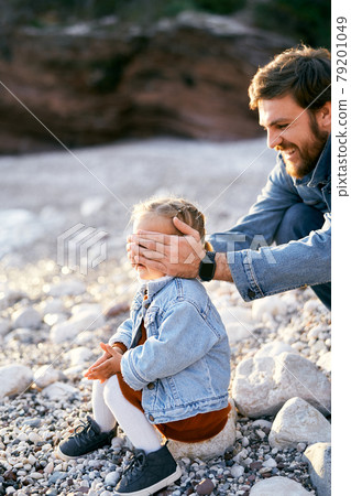 Smiling dad closed eyes of the little girl sitting on a stone in front of him with his palms Smiling dad closed eyes of the little girl sitting on a stone in front of him with his palms 79201049