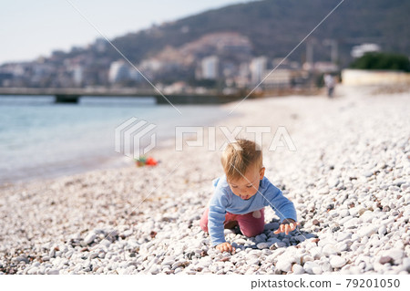 Pensive kid crawls along a pebble beach, sticking out his tongue, by the sea against the background of mountains Pensive kid crawls along a pebble beach, sticking out his tongue, by the sea against the background of mountains 79201050