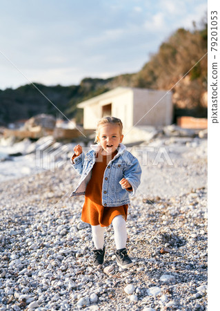 Little smiling girl in a dress and denim jacket walks along the pebble beach against the backdrop of the sea, rocks and building Little smiling girl in a dress and denim jacket walks along the pebble beach against the backdrop of the sea, rocks and building 79201053