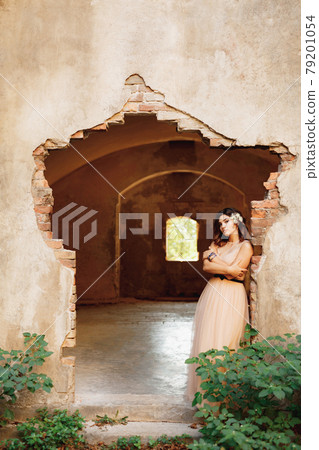 Lovely bride in a beautiful pastel dress with her arms crossed on her chest is pensively standing on the ruins of a castle 79201054