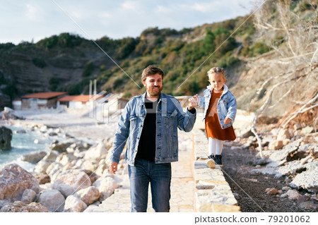 Smiling dad leads a little girl by the hand along the curb on a pebble beach against a background of green mountains. Close-up Smiling dad leads a little girl by the hand along the curb on a pebble beach against a background of green mountains. Close-up 79201062