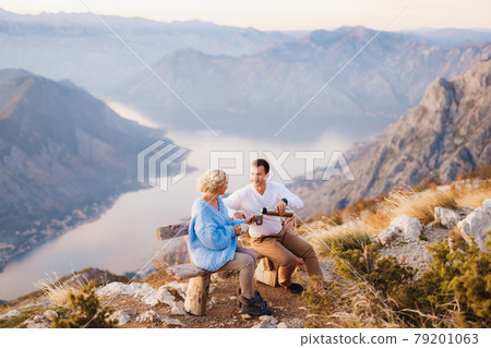 Couple sits on a bench and drinks tea against the backdrop of Mount Lovcen and the Bay of Kotor Couple sits on a bench and drinks tea against the backdrop of Mount Lovcen and the Bay of Kotor 79201063