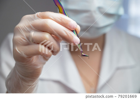 Lashmaker in a mask holds a bunch of eyelashes with tweezers. Close-up of a working tool of a master in a beauty salon Lashmaker in a mask holds a bunch of eyelashes with tweezers. Close-up of a working tool of a master in a beauty salon 79201669