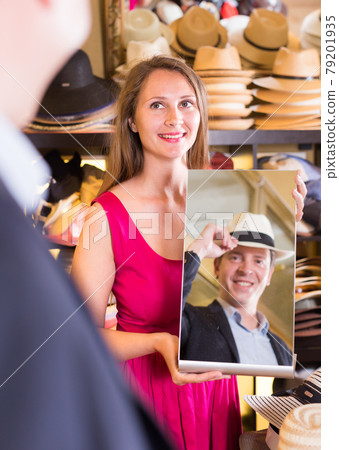 woman holding mirror and showing customer his reflection in hats shop 79201935