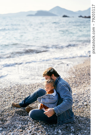 Dad in a denim jacket sits on a pebble beach and holds a little girl in his lap. Side view 79203117