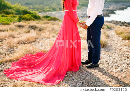 The bride in a long bright pink dress and the groom stand on a summer day in nature and tenderly hold each other's hands  79203120