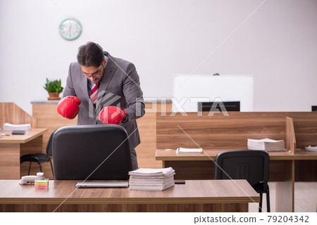 Young male employee wearing boxing gloves at workplace Young male employee wearing boxing gloves at workplace 79204342