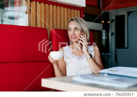 An attractive girl is talking on a mobile phone and smiling while sitting alone in a cafe 79205957