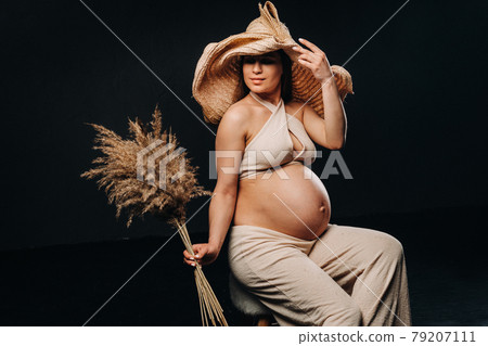pregnant woman in a straw hat with beige clothes with a bouquet in her hands in the studio on a black background 79207111