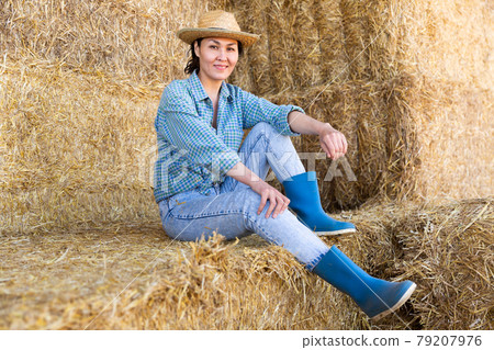 Positive relaxed asian female farmer resting on hayloft after work at farm, sitting on straw stack and smiling 79207976