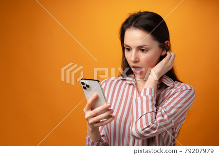 Surprised young woman using smartphone against yellow background 79207979