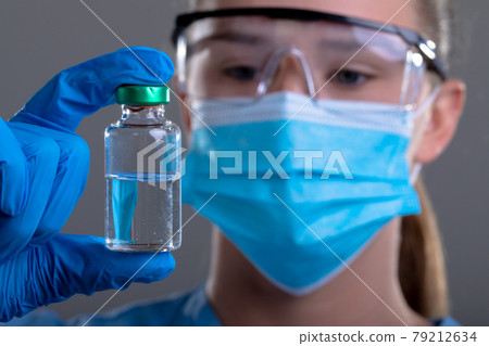 Caucasian female doctor wearing face mask and safety glasses holding vaccine vial 79212634