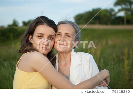 Senior mother with gray hair with her adult daughter looking at the camera in the garden and hugging each other during sunny day outdoors 79216280