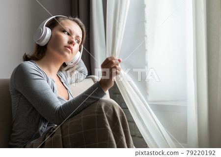 Portrait of young woman in headphones listening music sitting on sofa covered blanket and looking at window opening curtain Portrait of young woman in headphones listening music sitting on sofa covered blanket and looking at window opening curtain 79220192