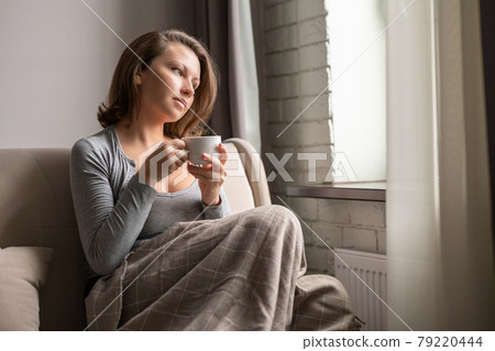 Portrait of beautiful young woman sitting on sofa covered blanket, drinking hot coffee from cup and looking at window. 79220444
