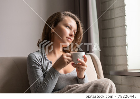 Portrait of beautiful young woman sitting on sofa covered blanket, drinking hot coffee from cup and looking at window. 79220445