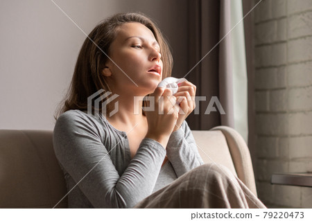 Sick brunette woman sneezing in a tissue sitting on the couch in the living room near window Sick brunette woman sneezing in a tissue sitting on the couch in the living room near window 79220473