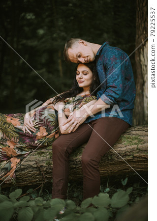 Young happy romantic pregnant couple sitting on the fallen tree in forest, hugging, touching belly 79225537