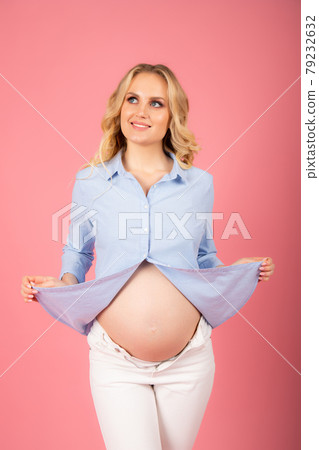 Beautiful pregnant woman in a blue shirt on a pink background. Naked belly 79232632