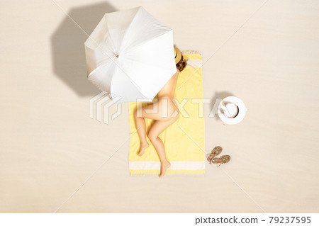 Alone naked woman in straw hat lying and sleeping on yellow beach towel under beach umbrella. Female relaxation on the sand beach at summer vacation. Top, aerial view 79237595