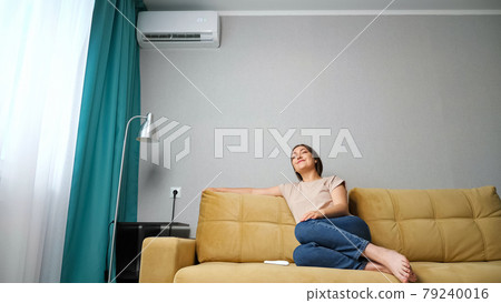 woman adjusts the air conditioner while sitting on the couch, woman adjusts the air conditioner while sitting on the couch, 79240016