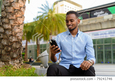Portrait of handsome black African businessman outdoors in city during summer sitting and using mobile phone while smiling 79241746