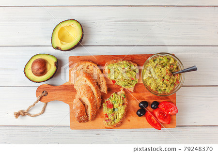 Flat lay photo, freshly prepared guacamole in small glass bowl, bread, tomatoes, olives at working board and two avocados next white wood desk 79248370