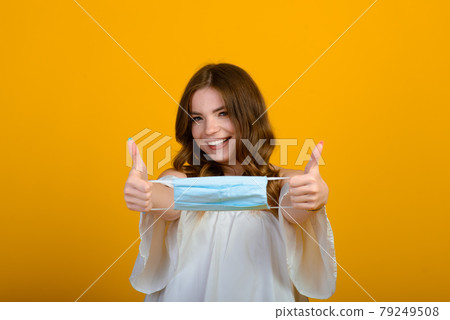 Beautiful happy young woman with face mask in hands on yellow background. COVID virus and air pollution. Recovering from illness, end of epidemic. Beautiful happy young woman with face mask in hands on yellow background. COVID virus and air pollution. Recovering from illness, end of epidemic. 79249508