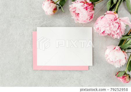Mockup invitation, blank paper greeting card, pink envelope and peonies on gray stone table. Flower background. Flat lay, top view. 79252958