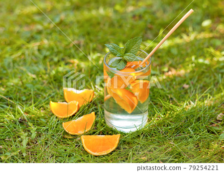 cold lemonade drink with orange and mint in tall glass on green grass and eco straw. glass with oranges and water on the grass 79254221