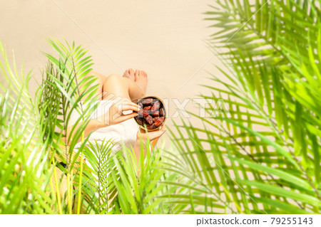 Woman in a straw hat sitting under coconut palm tree branches holding in a hand royal dates fruit in a bowl on the sand of beach. Summer. Top view 79255143