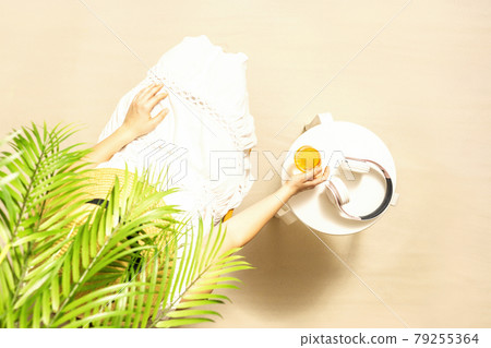 Alone woman sitting under palm tree branches. Glass of orange juice and headphones on the table. Female relaxation on the sandy beach at summer vacation. Top view 79255364