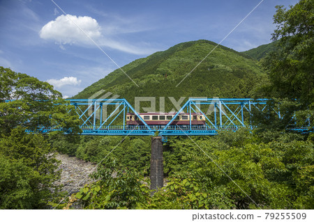 A one-car train crossing a light blue iron bridge under a blue sky with white clouds 79255509