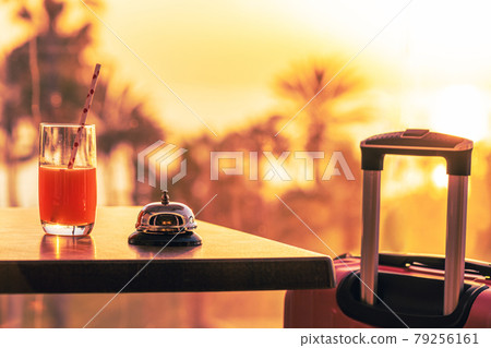Suitcase, ringing hotel service bell with welcome drink and sea, palm tree view on sunset. Travel concept. 24-hour hotel front desk. Late check-out. 79256161