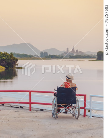 Back view of Asian elderly female patient wearing surgical mask and sitting on wheelchair with feeling lonely and depressed at riverside in the evening with view of Wat Tham Sua in background. Back view of Asian elderly female patient wearing surgical mask and sitting on wheelchair with feeling lonely and depressed at riverside in the evening with view of Wat Tham Sua in background. 79257682
