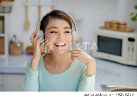 Attractive young woman listening to music with mobile phone and headphones while having breakfast in the kitchen 79269259
