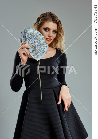 Blonde girl in black dress is holding some cash, posing against gray background. Gambling entertainment, poker, casino. Close-up. Blonde girl in black dress is holding some cash, posing against gray background. Gambling entertainment, poker, casino. Close-up. 79275542